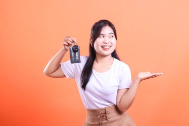 Neşeli genç bir kadın arabanın anahtarlarını tutuyor ve eliyle bir şey sunuyor, gülümsüyor. turuncu arkaplanda izole