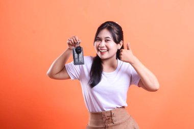 Heyecanlı genç bir kadın arabanın anahtarlarını elinde tutuyor, başparmakları havada ve gülümsüyor. Yeni araba almaktan mutlu, turuncu arka planda izole edilmiş..