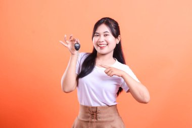 Mutlu genç Asyalı kadın yeni araba anahtarını tutuyor ve kameraya bakarak onu işaret ediyor. turuncu arkaplanda izole