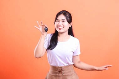 Asyalı bir kadın elinde mutlu bir gülümsemeyle araba anahtarı tutuyor. Sahiplik ya da yeni bir satın alma öneriyor. turuncu arkaplanda izole.