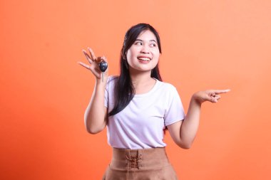 Elinde araba anahtarı olan genç Asyalı kadın, turuncu arka planda izole edilmiş sunum ya da sergi salonu teklifini gösteren bir tarafı mutlu bir şekilde işaret ediyor.