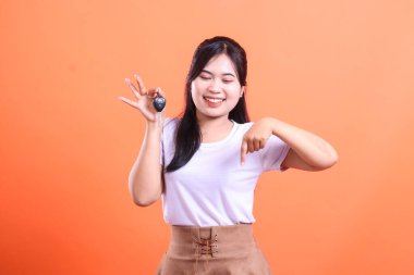 Neşeli bir Asyalı kadın arabanın anahtarlarını tutuyor ve gülümsüyor, arabanın anahtarına bakıyor ve turuncu arka planda izole edilmiş ürünü göstermek için parmağını aşağıya doğrultuyor.