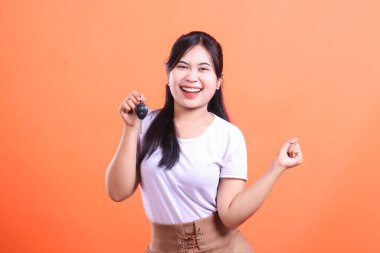 Gülümseyen ve elinde araba anahtarı olan neşeli, genç bir Asyalı kadın turuncu arka planda izole edilmiş yeni arabası için heyecanlı görünüyor..