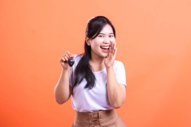 Güzel, heyecanlı Asyalı bir kadın araba anahtarlarını mutlu bir ifadeyle gösteriyor ve eli ağzının kenarında, başarıyı temsil ediyor. turuncu arkaplanda izole