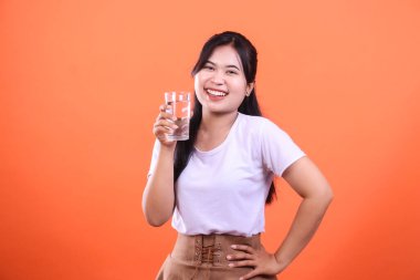 Elinde bir bardak su tutan, sağlıklı bir yaşam tarzını destekleyen ve turuncu arka planda günlük olarak susuz kalan genç bir kadın..