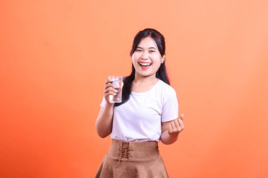 Gülümseyen genç bir kadın elinde neşeli bir ifadeyle bir bardak su tutuyor. Turuncu arka planda izole edilmiş sağlıklı su alışkanlıklarını teşvik ediyor..