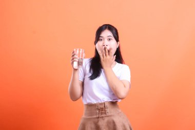 Genç Asyalı bir kadın, turuncu arka planda izole edilmiş bir şekilde elini bir bardak suya uzatarak şok geçiriyor..