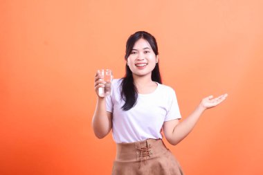 Gülümseyen genç kadın elinde bir bardak temiz su tutuyor, susuzluğu ve sağlıklı yaşam tarzını destekliyor. turuncu arkaplanda izole.