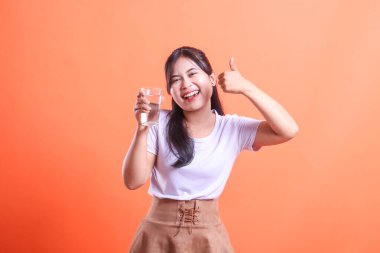 Neşeli genç bir kadın elinde bir bardak su tutuyor ve iyi niyetli bir jest yapıyor. Susuzluğu ve sağlıklı bir yaşam tarzını teşvik ediyor. turuncu arkaplanda izole