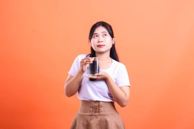 Beyaz tişörtlü genç bir kadın bir bardak su tutuyor ve yüzünde düşünceli bir ifadeyle yukarı bakıyor. turuncu arkaplanda izole