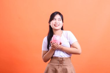 Genç Asyalı kadın elinde pembe bir kumbara tutuyor. Finansal planlamayı ve gelecek için biriktirmeyi simgeliyor. turuncu arkaplanda izole