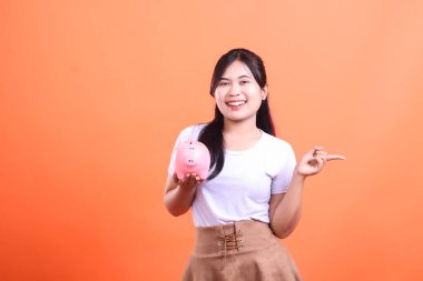 Pembe domuz kumbarası tutan neşeli Asyalı genç bir kadın ve bir kenarı göstererek, fotokopi alanı gösteriyor. turuncu arkaplanda izole.