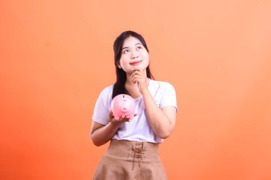 Asyalı bir kadın elinde kumbara tutarken yukarı bakıyor ve gülümsüyor. Turuncu arka planda izole edilmiş para biriktirme kavramını temsil ediyor.