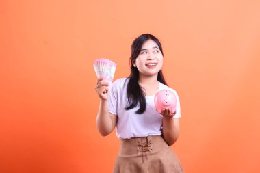 Elinde banknotlar ve domuz kumbarası olan gülümseyen genç bir kadın gözlerini kaçırırken hayal kurarak turuncu arka planda izole edilmiş birikimini düşünüyor..