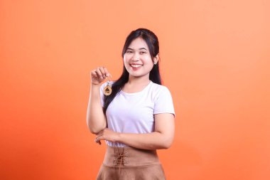 Genç bir kadın elinde ev cazibesiyle anahtarlarını tutarken gülümsüyor. Yeni ev alımı ve gayrimenkul başarısı için heyecanlanıyor. turuncu arkaplanda izole