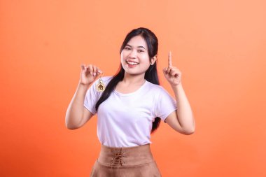 Genç bir kadın gülümsüyor ve elinde küçük bir ev kolyesi var. Parmağıyla yukarıyı gösteriyor. Turuncu arka planda izole edilmiş yeni ev sahipleri için neşeli bir an.