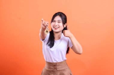 Genç Asyalı kadın ev anahtarlarını tutuyor ve onay veriyor, yeni ev alımını kutluyor, turuncu arka planda izole edilmiş.