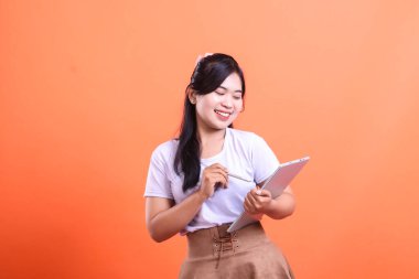 Stüdyo çekimlerinde gülümserken tablet kullanan genç ve güzel bir Asyalı kadın. Elinde bir stil vardı. turuncu arkaplanda izole