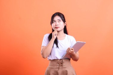 Düşünceli genç bir kadın elinde bir tablet tutuyor ve dikkatlice yukarı bakıyor. Beyaz tişört ve etek giyiyor, turuncu arka planda izole edilmiş..