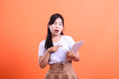 Şaşırmış Asyalı kadın şok olmuş bir ifadeyle tablet tutuyor ve ekranı parmağıyla işaret ediyor. turuncu arkaplanda izole.