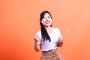 Heyecanlı genç kadın bir tablet ve kalem tutuyor, neşe saçıyor ve çabalarında başarıya ulaşıyor. turuncu arkaplanda izole.