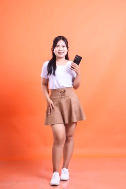 Genç Asyalı bir kadın elinde akıllı bir telefonla gülümseyerek poz veriyor ve gelişigüzel giyiniyor. turuncu arkaplanda izole.
