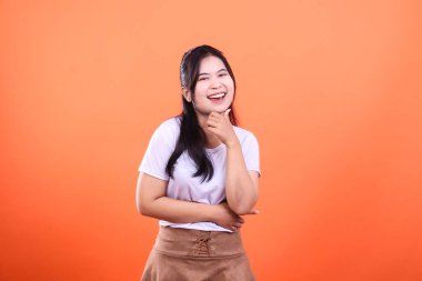 Siyah saçlı, beyaz tişörtlü ve kahverengi etekli bir kadın eli çenesinde turuncu bir arka planda poz veriyor.