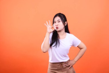 Uzun siyah saçlı genç bir kadın beyaz bir gömlek ve kahverengi bir etek giyiyor. İyi bir el hareketi ve turuncu bir arka planda izole edilmiş dudaklarla poz veriyor..