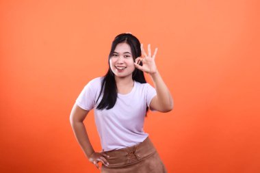 Uzun siyah saçlı bir kadın gülümserken iyi bir el hareketi sergiliyor, turuncu arka planda izole edilmiş, beyaz tişört ve kahverengi etek giyiyor, resim stüdyo fotoğrafı..