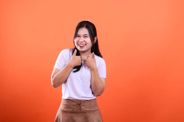 Beyaz gömlekli, kahverengi etekli neşeli bir kadın gülümser ve turuncu bir arka planda, göğsünün önünde iki baş parmağını kaldırır.