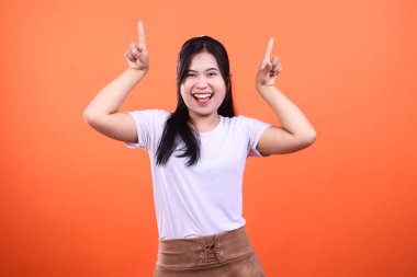 Beyaz gömlek ve kahverengi etek giymiş, kameraya mutlu bir şekilde gülümseyen bir kadın turuncu arka planda izole bir şekilde duruyor ve işaret parmaklarının ikisini de işaret ediyor..