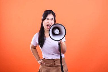 Bir kadın, turuncu arka planda izole bir şekilde megafona bağırıyor. Beyaz bir gömlek ve kahverengi bir etek giyiyor..