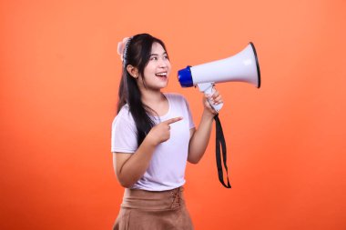 At kuyruklu genç bir kadın beyaz ve mavi bir megafon tutuyor ve işaret parmağıyla işaret ediyor. Kadın beyaz bir tişört ve kahverengi bir etek giyiyor. Turuncu bir arka planda izole edilmiş..