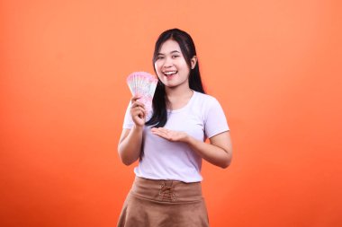 Elinde Endonezya Rupiah banknotları olan, beyaz gömlek ve kahverengi etekli, turuncu arka planda izole edilmiş bir kadın..