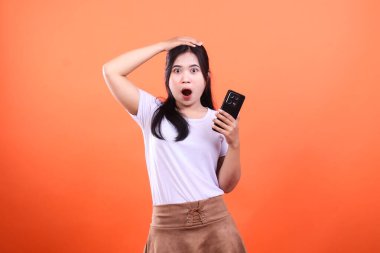 Şok olmuş bir ifadeyle genç bir kadın elinde akıllı bir telefon tutuyor. Bir eli kafasında, ağzı açık ve turuncu bir arka planda izole edilmiş..