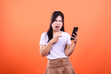 Beyaz tişört ve kahverengi etek giyen bir kişi bir elinde siyah bir akıllı telefonu tutarken diğer elinde turuncu bir arka planda izole edilmiş kızgın bir ifadeyle..
