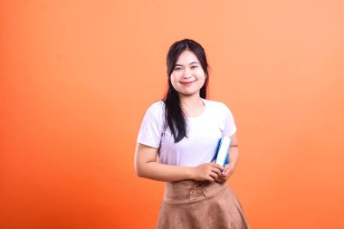 Bir kadın elinde mavi bir kitapla turuncu bir arka planda dikilip poz veriyor.