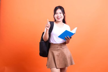 Sırtında siyah bir sırt çantasıyla elinde açık mavi bir kitapla gülümseyen bir insan, turuncu arka planda tek parmağını kaldırarak duruyor.