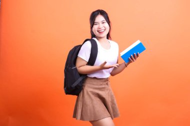 Siyah sırt çantalı genç bir kadın elinde bir kitapla turuncu arka planda poz veriyor..