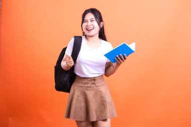 Siyah sırt çantalı genç bir kadın gülümserken mavi bir kitap tutuyor. Beyaz bir gömlek ve kahverengi bir etek giyiyor, turuncu arka planda izole edilmiş..