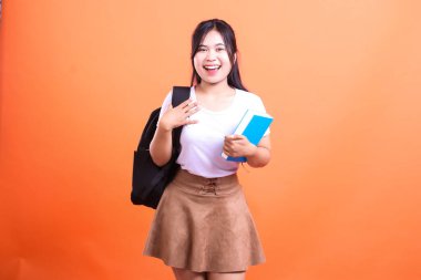Sırt çantası ve mavi kitabı olan gülümseyen bir kız öğrenci turuncu arka planda izole edilmiş bir stüdyoda poz veriyor..