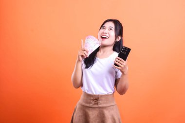 Genç bir kadın bir elinde para yelpazesi, diğer elinde ise turuncu arka planda izole edilmiş bir heyecan ifadesi olan akıllı bir telefon tutuyor..