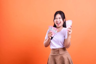 Bir kadın elinde iki deste Endonezya Rupiah banknotuyla mutlu bir ifadeyle gülümsüyor. Kadın beyaz bir bluz ve kahverengi bir etek giyiyor. turuncu bir arkaplanda izole