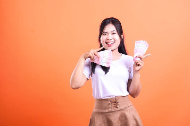 Bir kadın elinde birden fazla Endonezya Rupiah banknotu tutarken gülümsüyor. O, beyaz bir tişört ve kahverengi bir etek giyiyor. Arka plan turuncu..