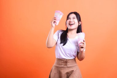 Bir kadın elinde iki deste rupiah para tutuyor ve gülüyor. Beyaz tişört ve kahverengi etek giyiyor, turuncu arka planda izole edilmiş..