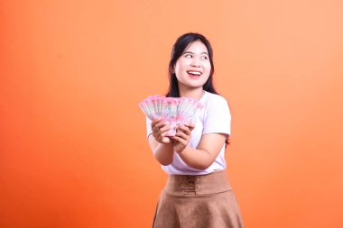 Elinde kağıt para tutan ve turuncu arka planı olan bir kadın. Kadın beyaz bir gömlek ve kahverengi bir etek giyiyor. turuncu arkaplanda izole