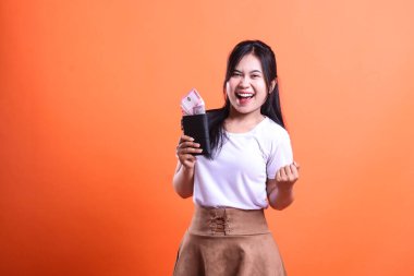 Elinde rupia parası olan siyah bir cüzdanı tutan genç bir kadın turuncu bir arka planda beyaz gömlek ve kahverengi etek giyiyor ve çok mutlu..