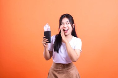 Endonezya Rupia banknotlarıyla dolu siyah bir cüzdan taşıyan bir kadın turuncu arka planda şaşırmış ve mutlu görünüyor.