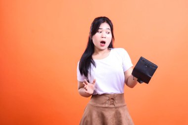 Şok olmuş bir kadın gözlerini kaçırırken boş bir siyah cüzdan tutuyor. O, beyaz bir gömlek ve kahverengi bir etek giydi. Arkaplan turuncu arka planda sade turuncu..