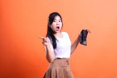 Boş cüzdanlı bir kadın şaşırmış ve solu işaret ediyor. turuncu arkaplanda izole